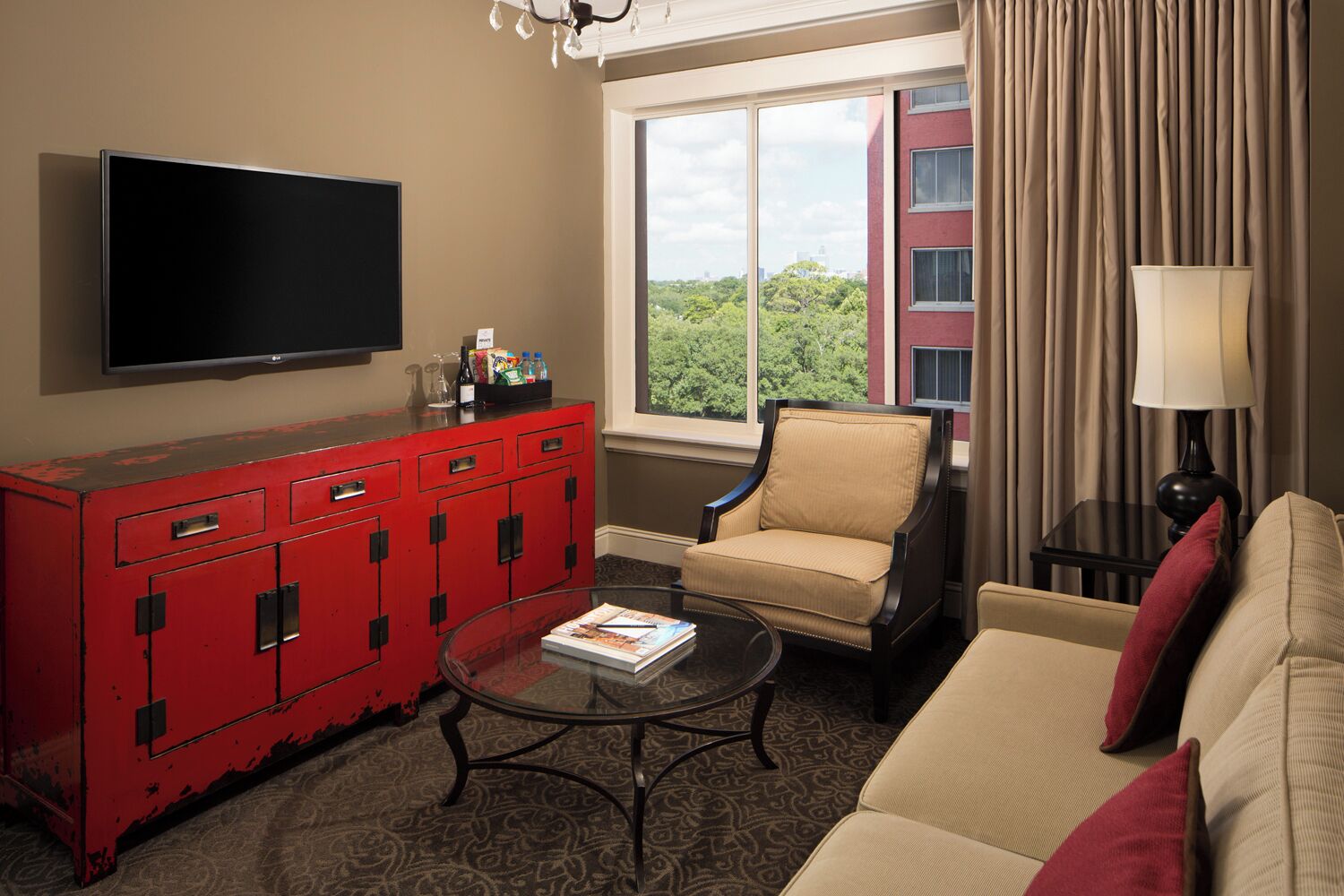 Habitación de hotel con televisión, un armario rojo, un sillón beige y una mesa de centro de vidrio, con vistas a zonas verdes.