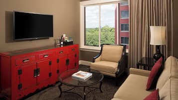Habitación de hotel con televisión, un armario rojo, un sillón beige y una mesa de centro de vidrio, con vistas a zonas verdes.