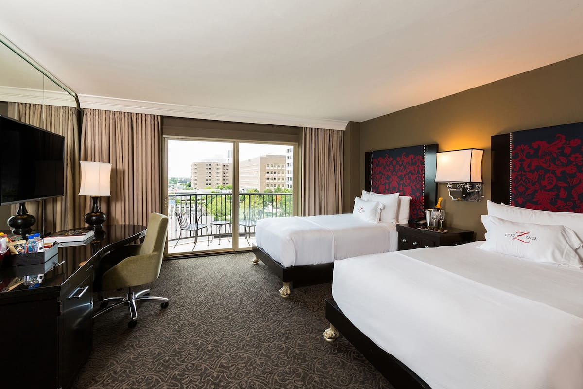 A hotel room with two beds, a desk, a chair, a TV, and a balcony with a view of buildings.