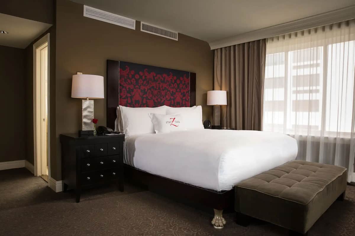 A hotel room with a large bed, two bedside lamps, a dark dresser, a decorative headboard, and a window with curtains.