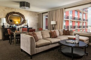 Habitación de hotel con sofá color beige, una mesa de comedor, una mesa de centro, un espejo grande y vista a un edificio rojo con balcones.