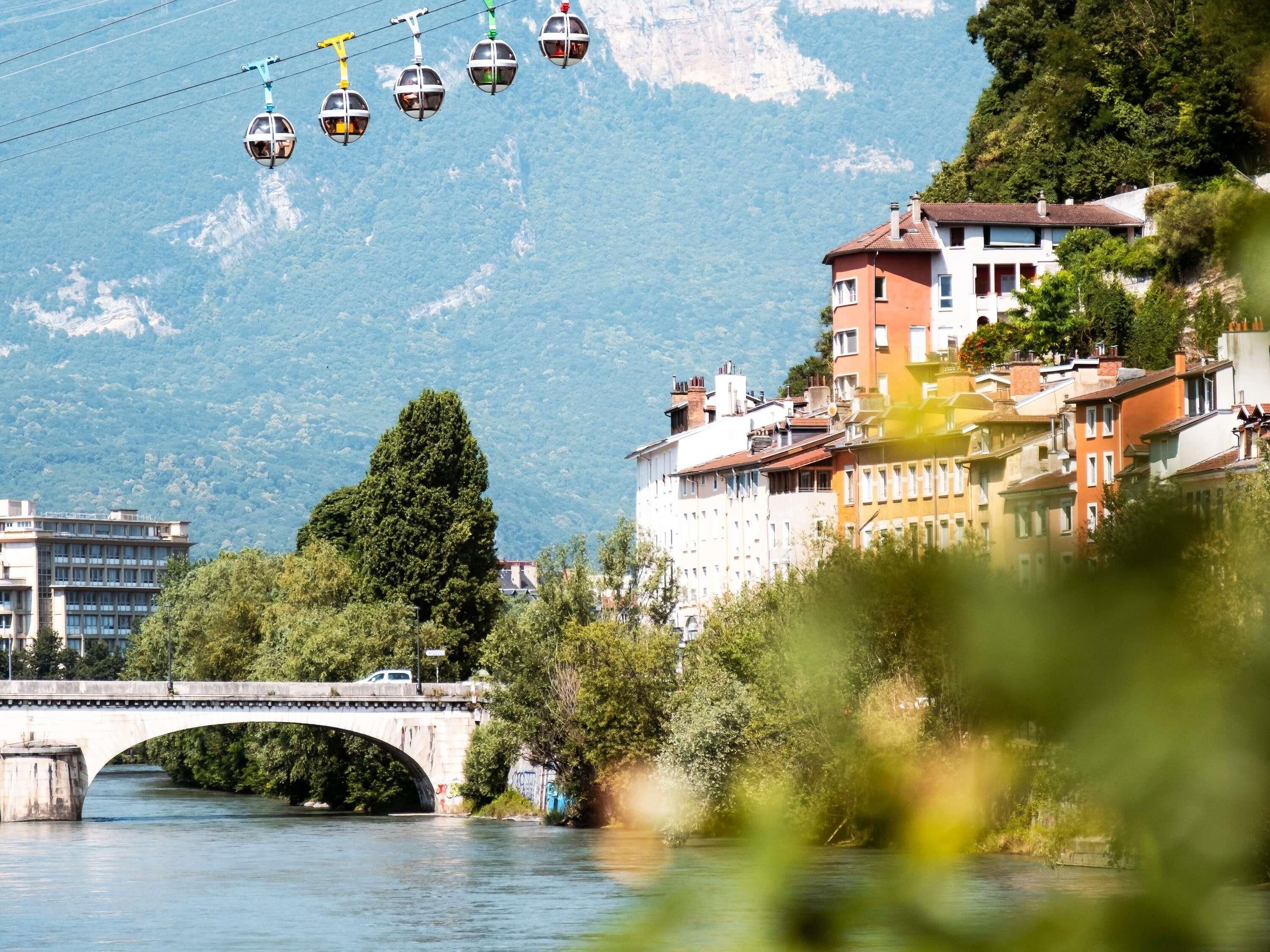 Eine Seilbahn, eine Brücke und Gebäude entlang eines Flusses mit Bergen im Hintergrund.