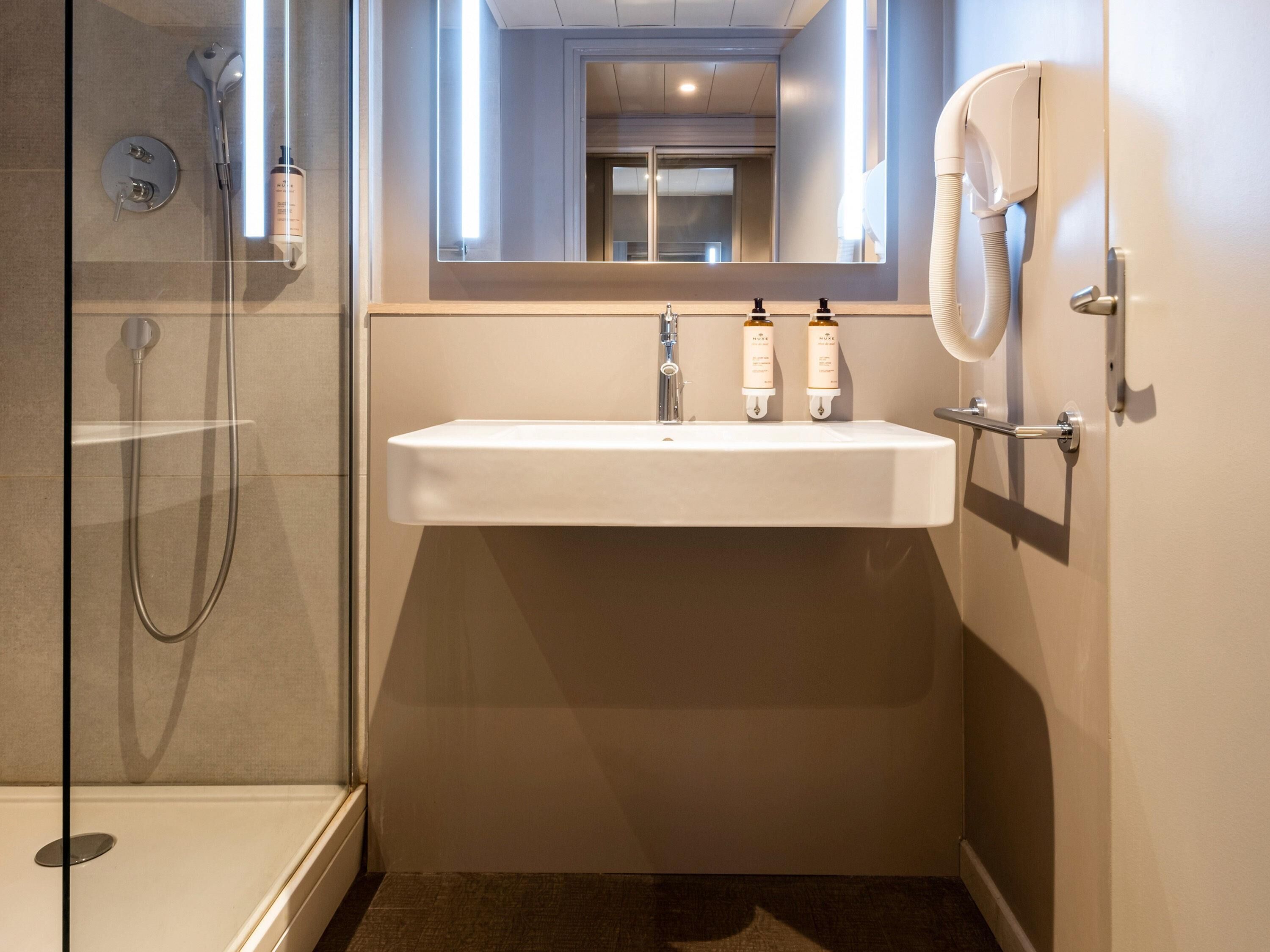 Un baño moderno con cabina de ducha de vidrio, un lavamanos blanco y dos botellas de champú, y un perchero para toallas.