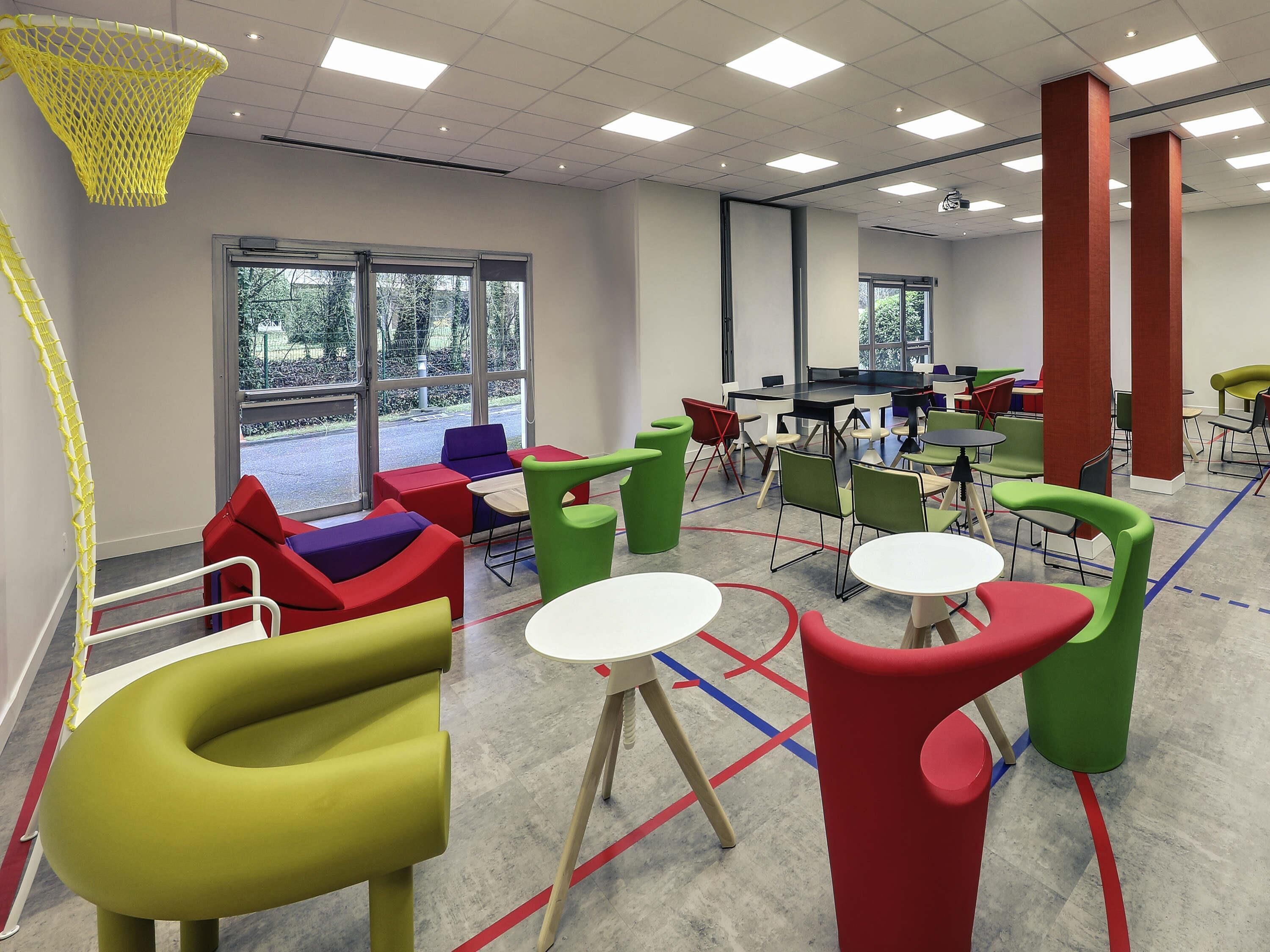 A brightly lit indoor space with colorful chairs and tables, large windows, and a basketball hoop.