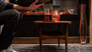 A person reading a book in front of a fireplace with a glass of red drink on a small wooden table.