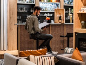 A person seated at a bar with a drink, bar shelves stocked with bottles, and a wicker chair in the foreground. - Mercure Grenoble Meylan (Meylan)