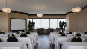 Sala de conferências com mesas brancas, cadeiras e um projetor.