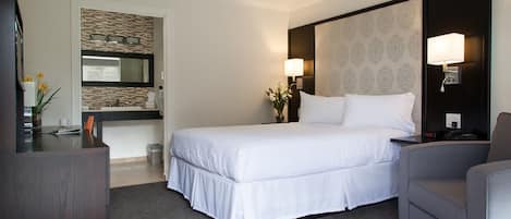 A hotel room with a large bed, a desk, a chair, a nightstand, a lamp, and a mirror.