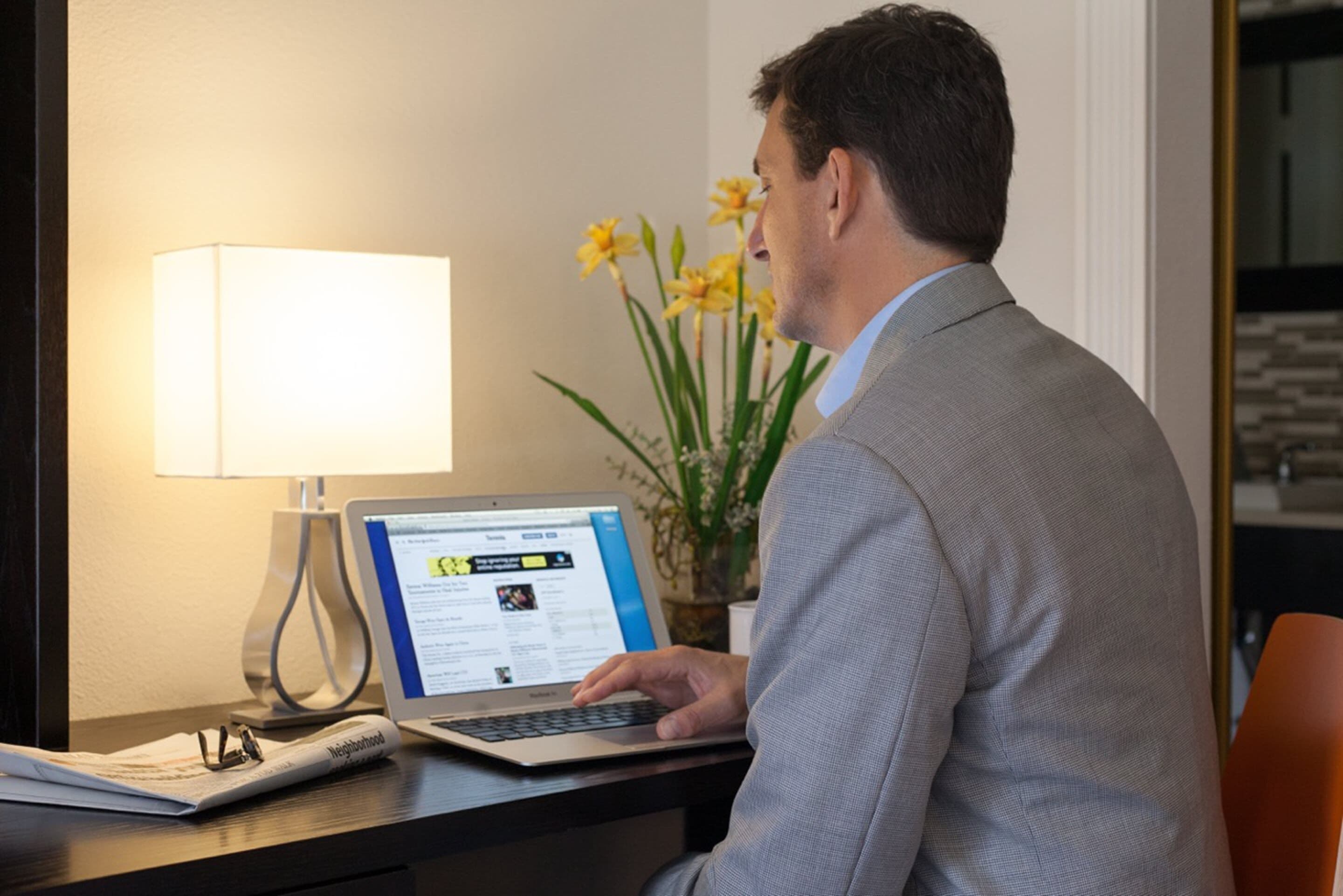 A man sitting at a desk with a laptop, a lamp, and a vase of flowers.