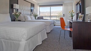 Quarto de hotel com duas camas, uma mesa e uma cadeira. Há uma janela grande com vista para o exterior.