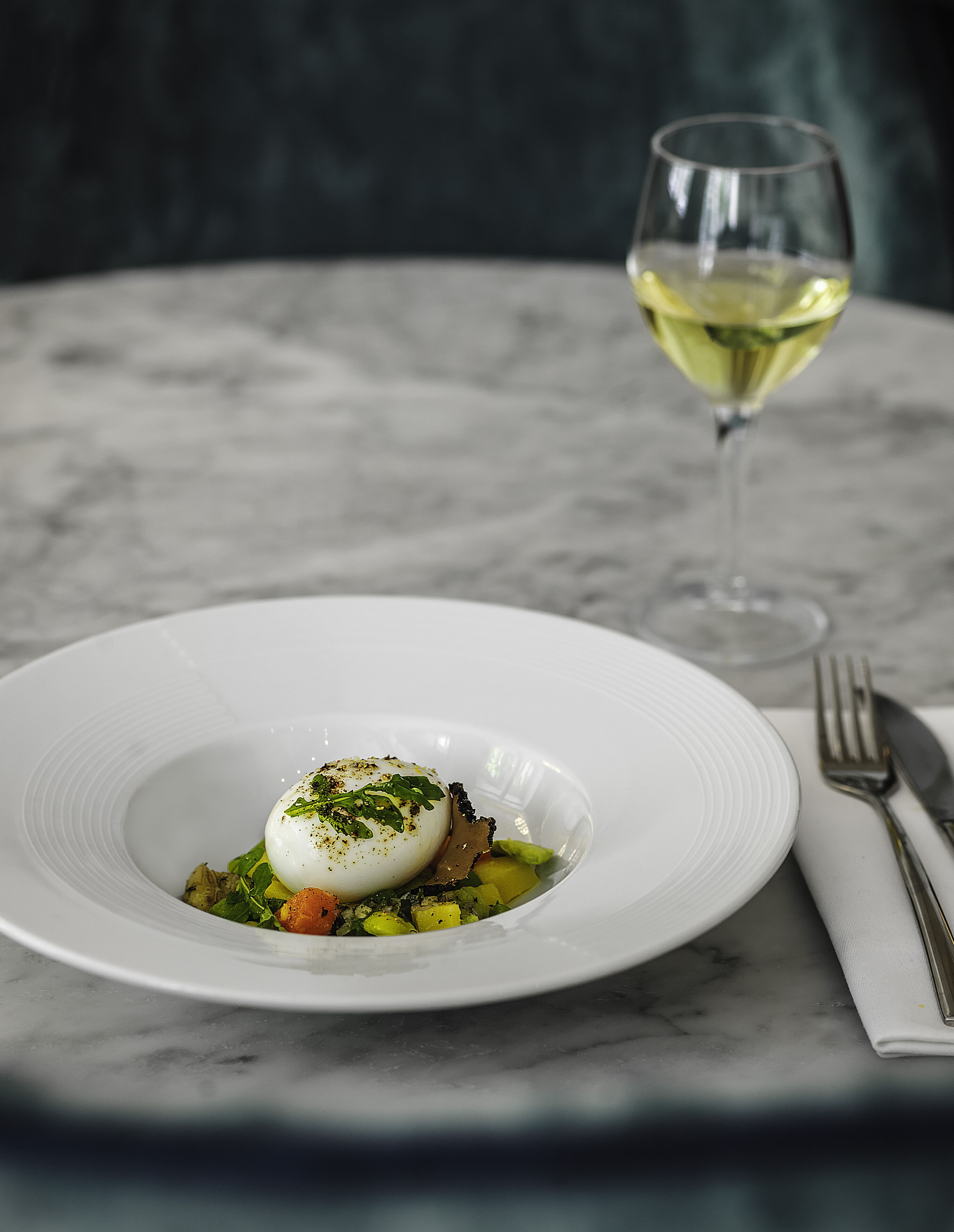 Une assiette blanche contenant un œuf poché, des légumes verts et d’autres légumes, posée sur une table en marbre. Un verre de vin blanc et des couverts se trouvent à l’arrière-plan.
