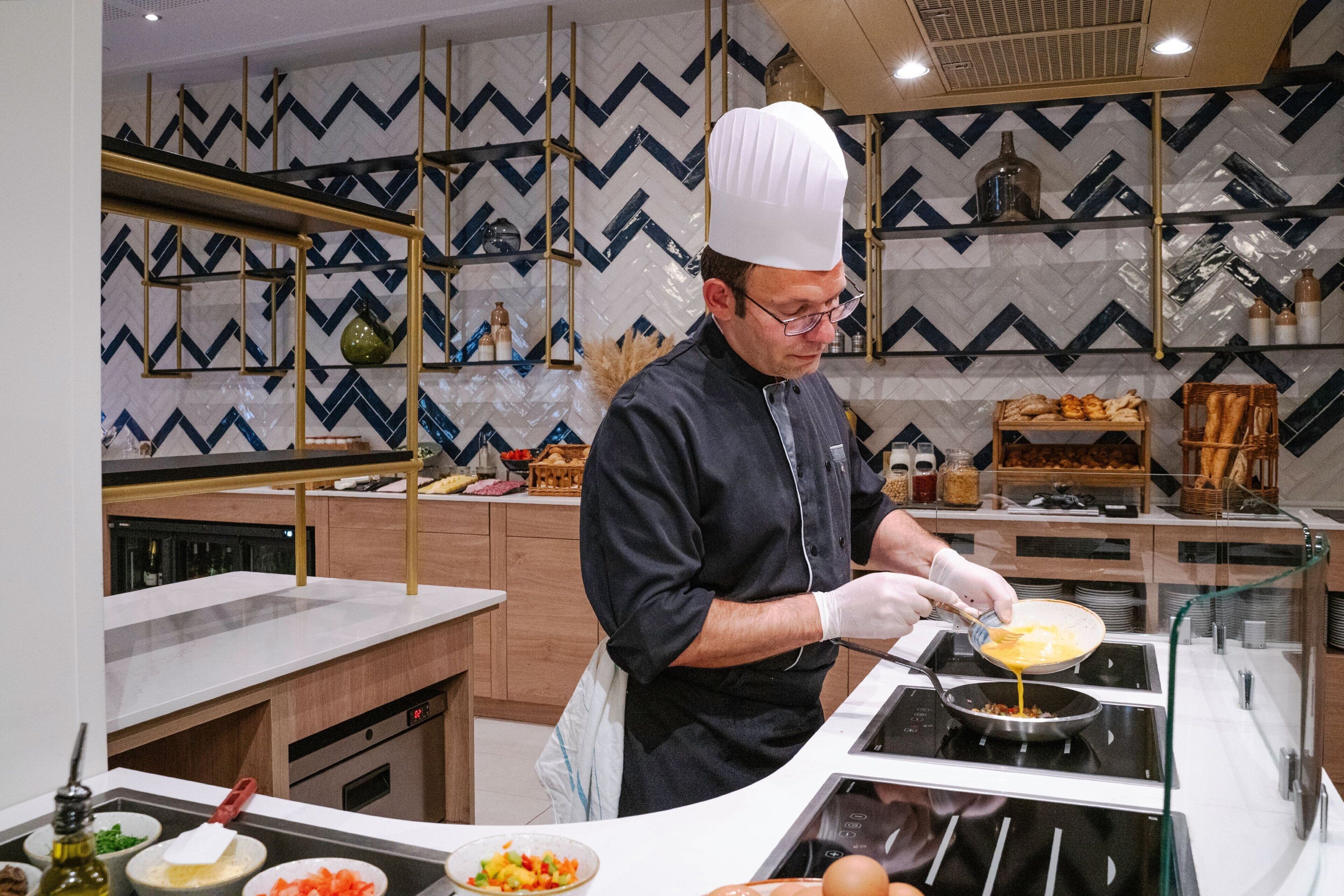 Uno chef in una cucina moderna con piastrelle a motivi geometrici, elettrodomestici in acciaio inossidabile ed scaffali in legno.