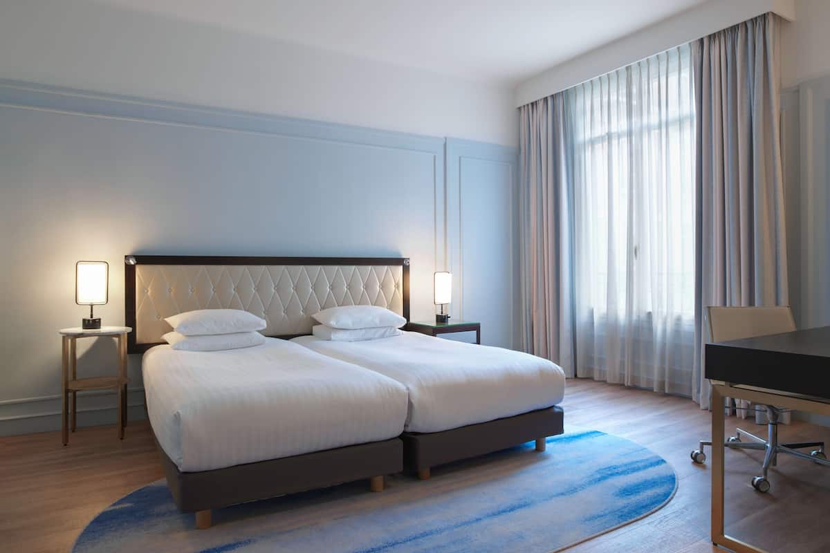 A double bed with a tufted headboard, two bedside tables with lamps, a desk with a chair, and a large window with curtains.
