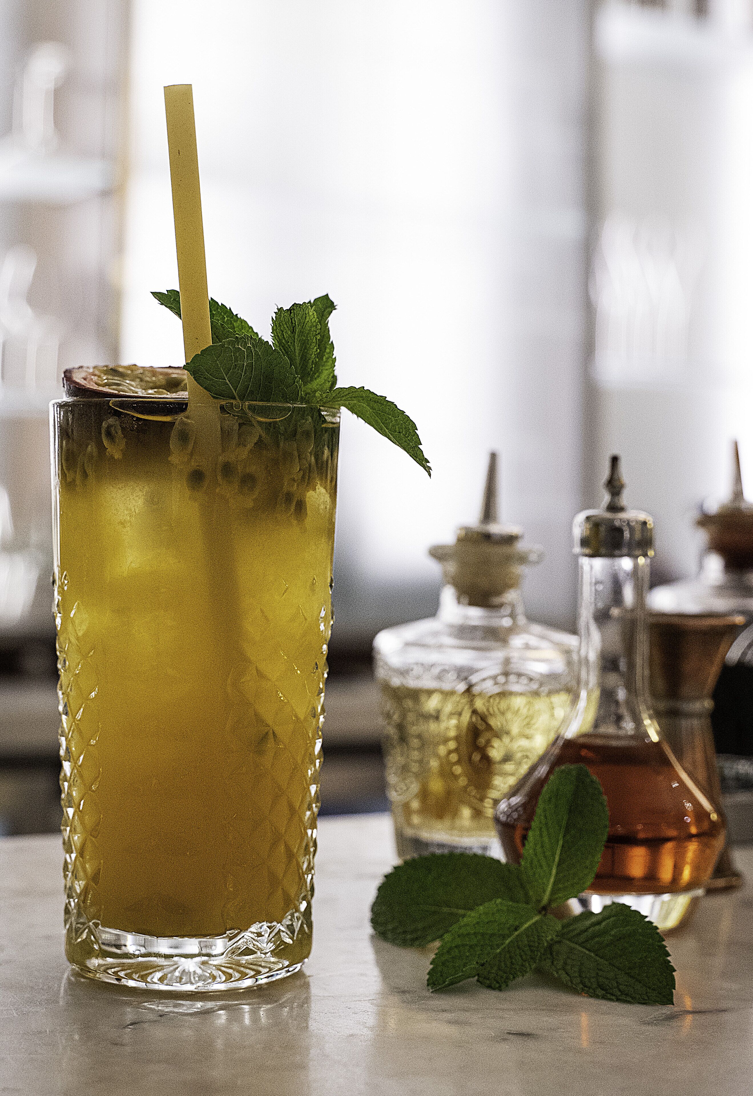 Un vaso de bebida amarilla con pajita y hojas de menta, acompañado de dos botellas de perfume.