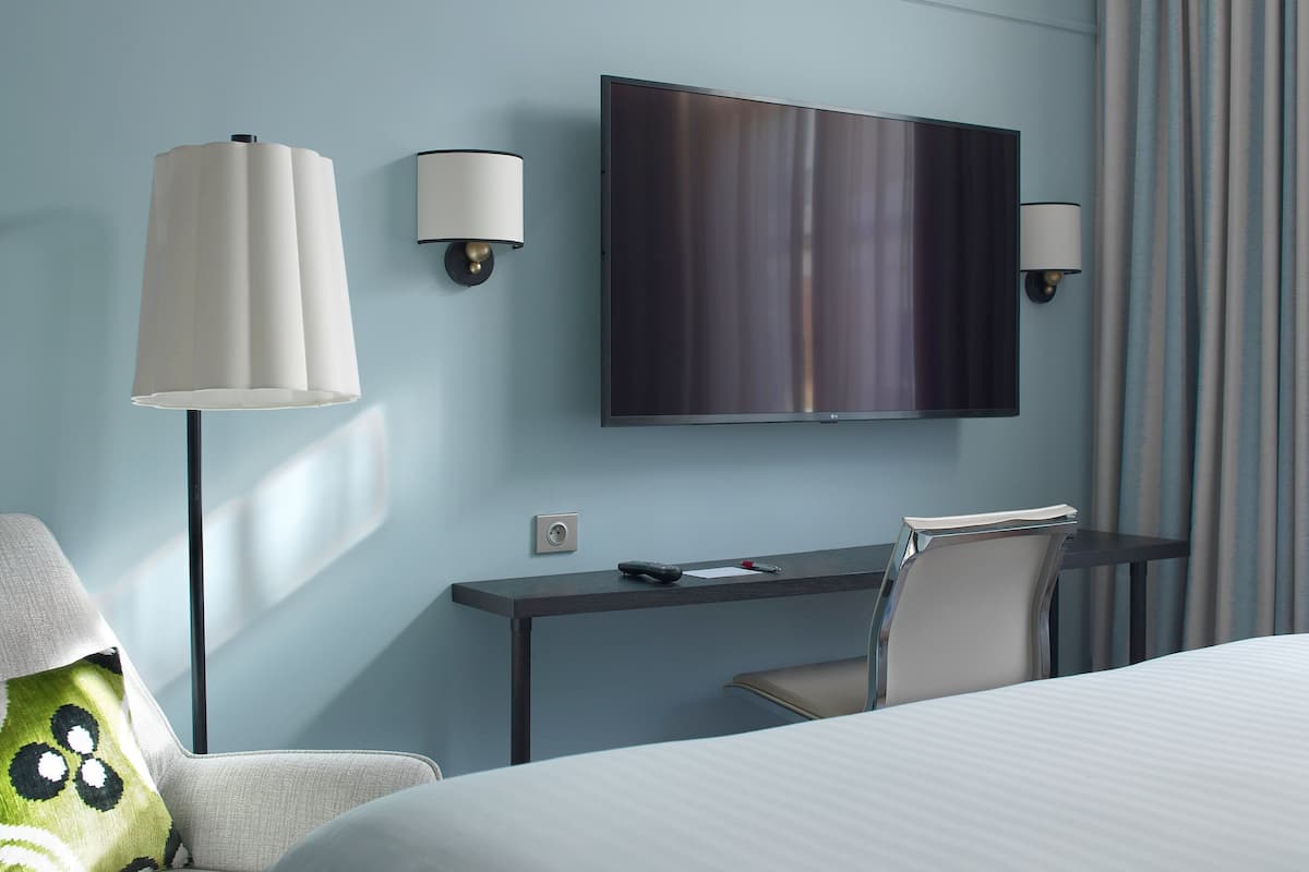 A hotel room with a bed, a desk, a chair, a TV, and two wall-mounted lamps.
