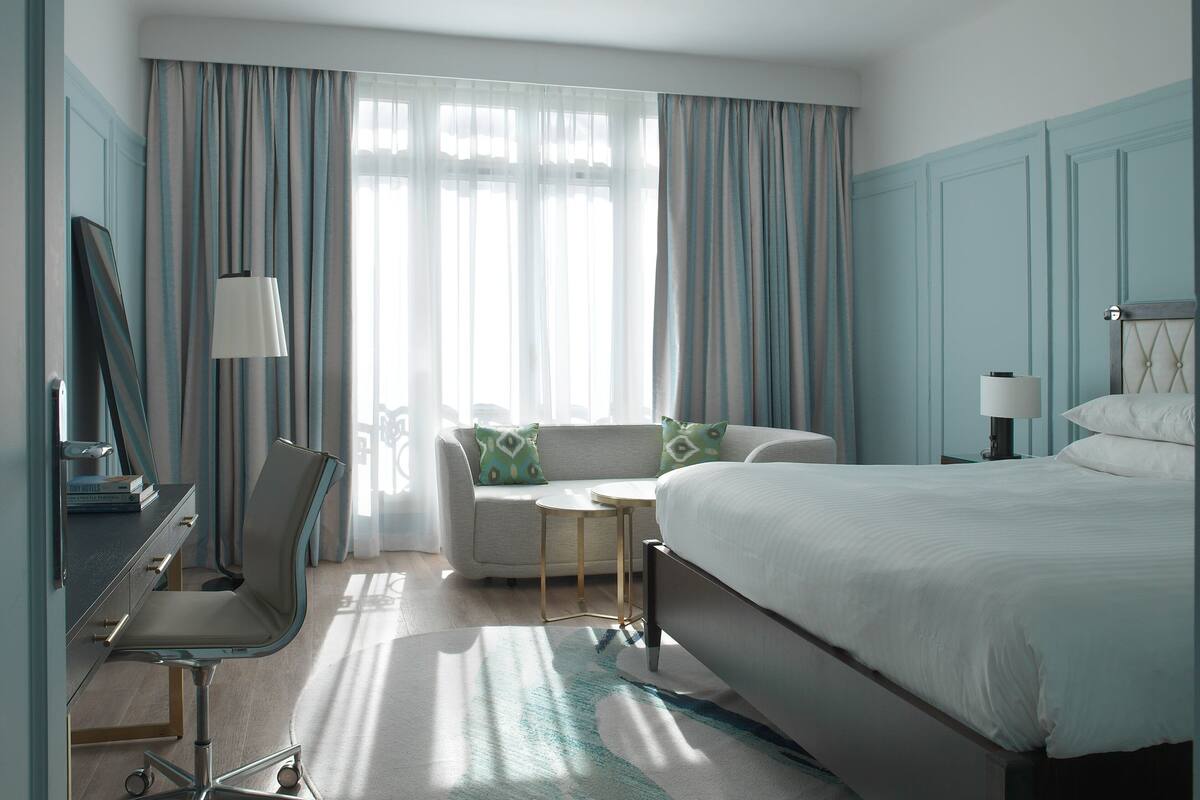 A hotel room with a bed, a desk with a chair, a small table, and a sofa with cushions.