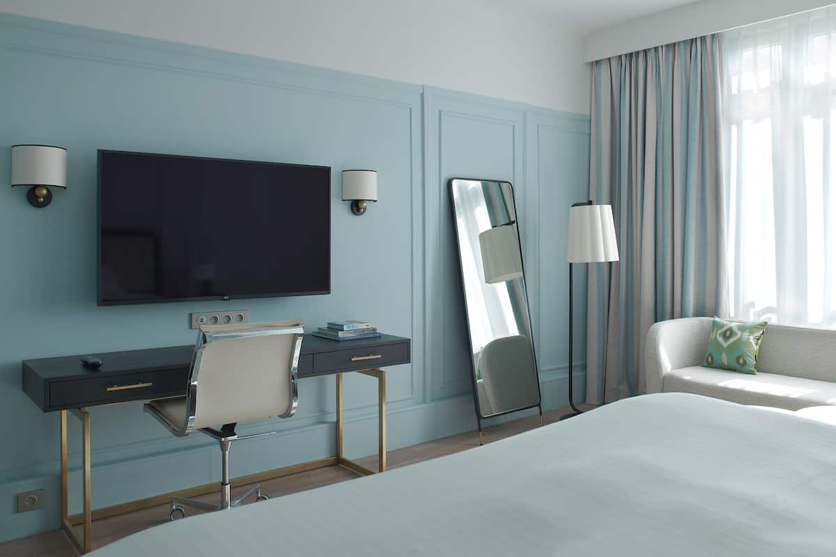 A hotel room with a bed, a desk with a chair, a flat-screen TV, a large mirror, and a sofa with a pillow.
