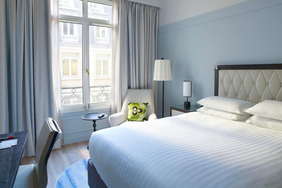 A hotel room with a bed, a chair, a lamp, a small table, and a window with curtains.