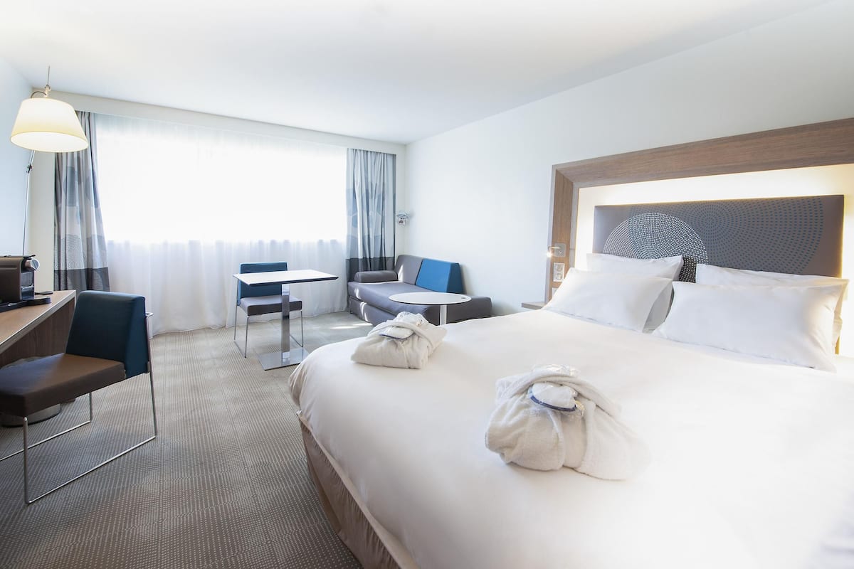 A hotel room with a large bed, a desk, a chair, a sofa, and a window with curtains.