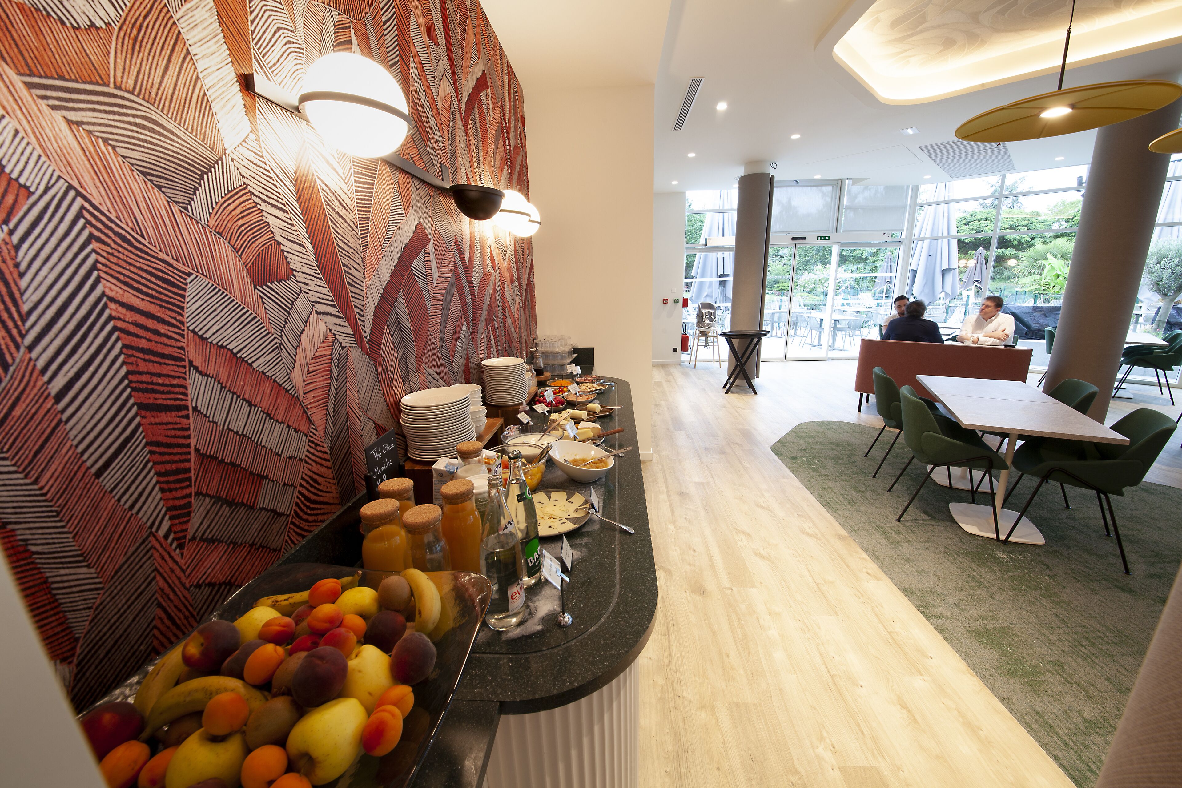Un área de bufé con una variedad de frutas y bebidas, un muro con diseño, y un espacio de estar con mesas y sillas.