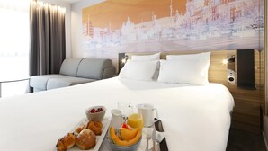 Una habitación de hotel con una cama, una bandeja de desayuno, un mural de un paisaje urbano y una ventana con cortinas.