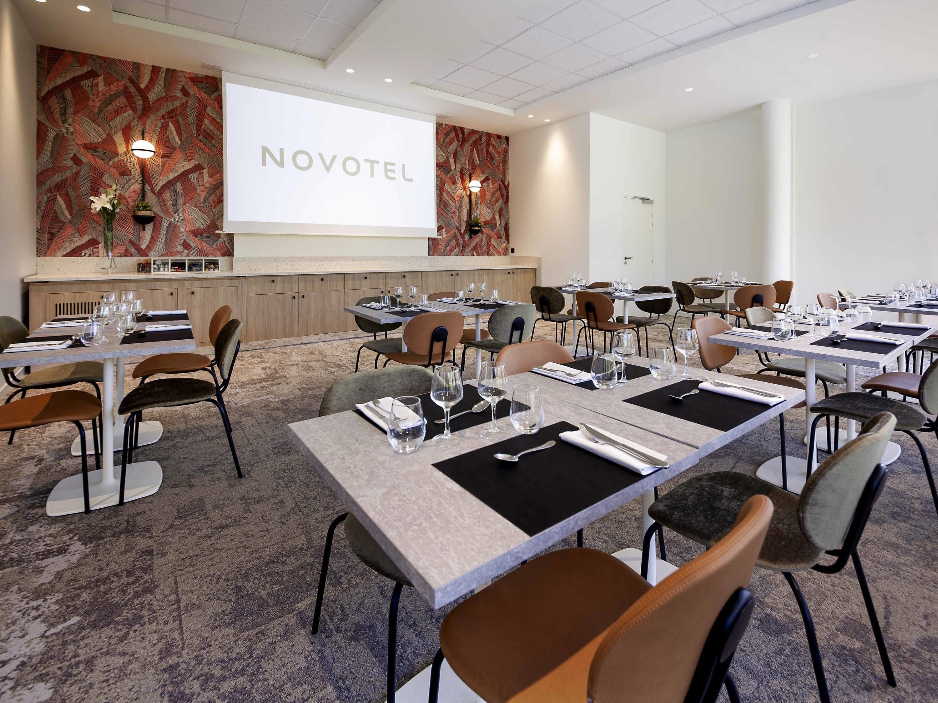Uma sala de conferências com mesas preparadas para uma reunião, uma tela de projeção exibindo "NOVOTEL" e uma parede decorativa com padrão de folhas.