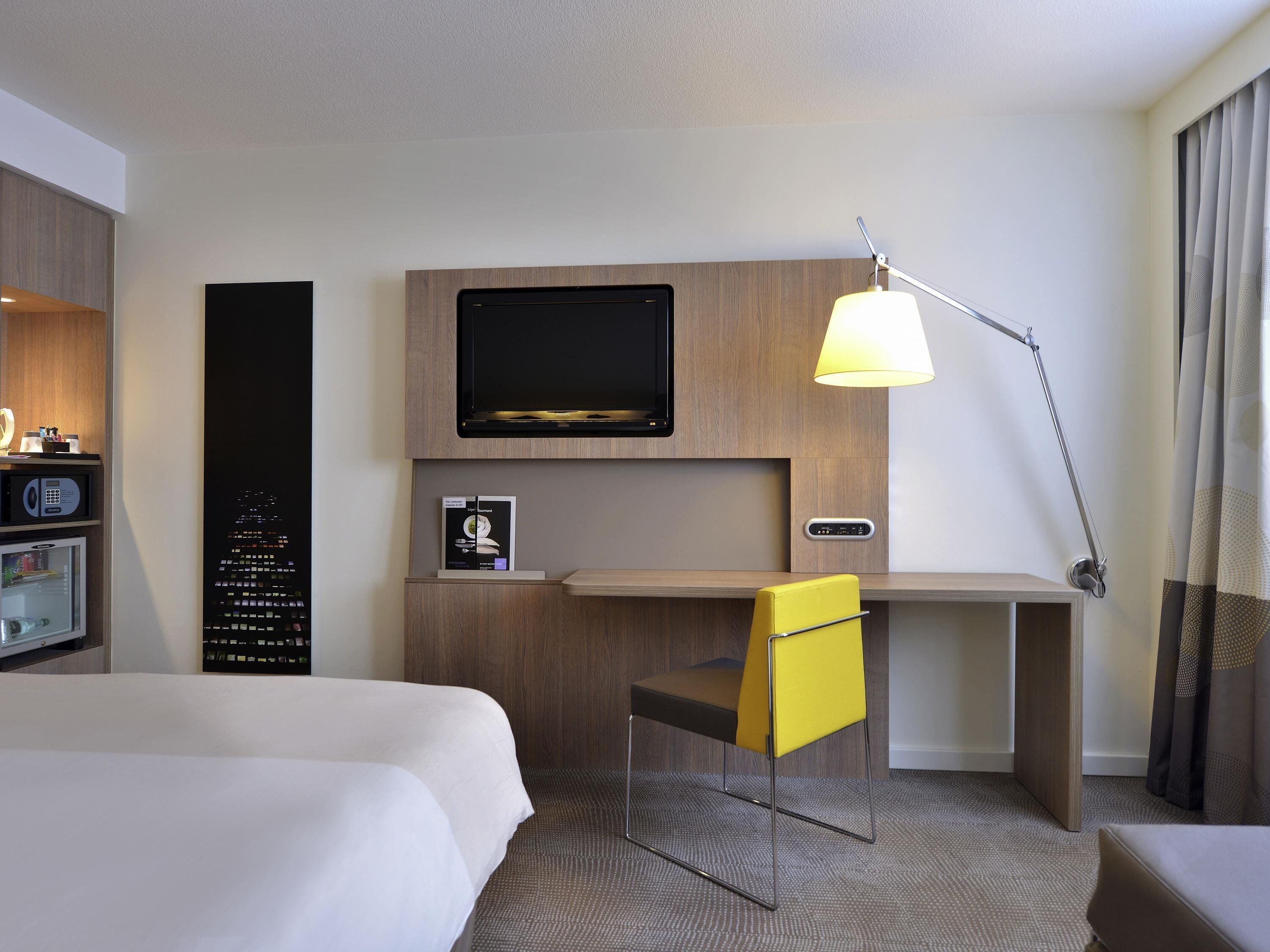 Una habitación de hotel con cama, escritorio, televisor, lámpara y minibar.