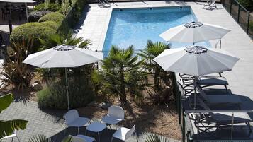 Piscina con sillas de descanso y sombrillas, rodeada de vegetación y un patio empedrado.
