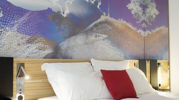 Habitación de hotel con un mural grande que muestra un paisaje nevado, una cama con almohadas blancas y rojas, y una mesita de noche con una lámpara.