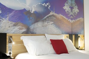 Habitación de hotel con un mural grande que muestra un paisaje nevado, una cama con almohadas blancas y rojas, y una mesita de noche con una lámpara.