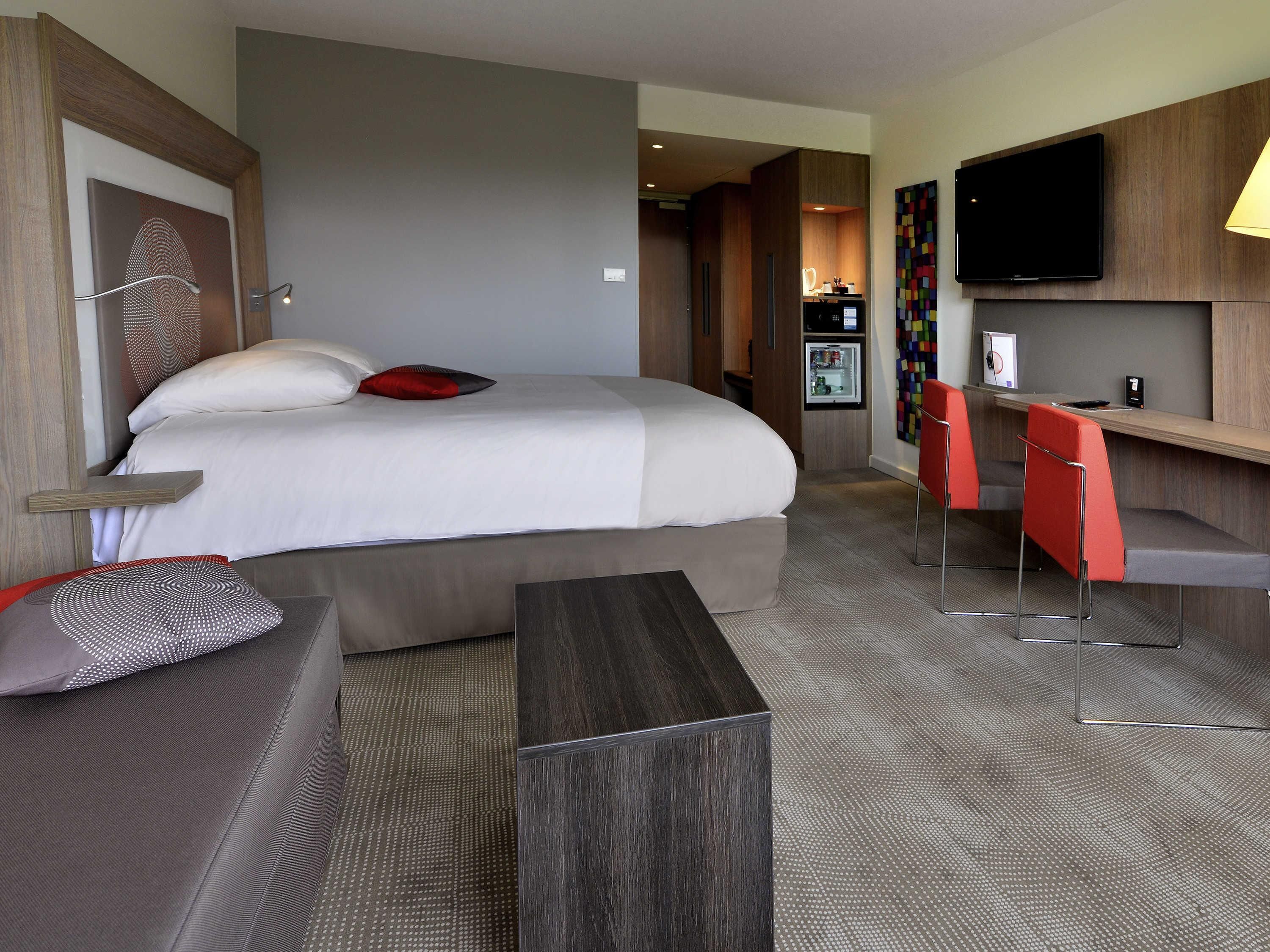 A hotel room with a bed, a desk, two chairs, a television, and a refrigerator.