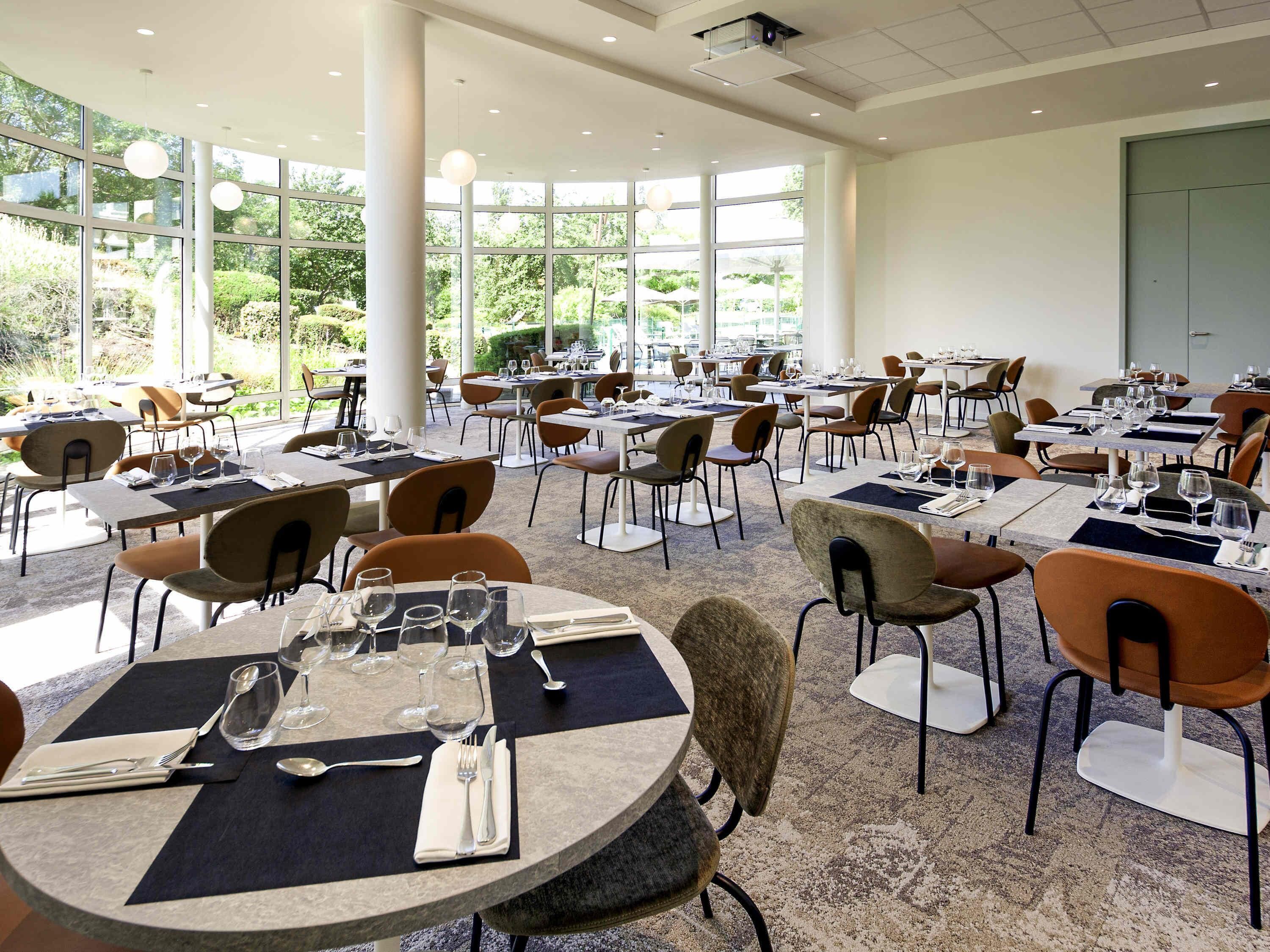 A spacious indoor dining area with multiple tables and chairs, large windows offering a view of greenery, and a modern ceiling design.