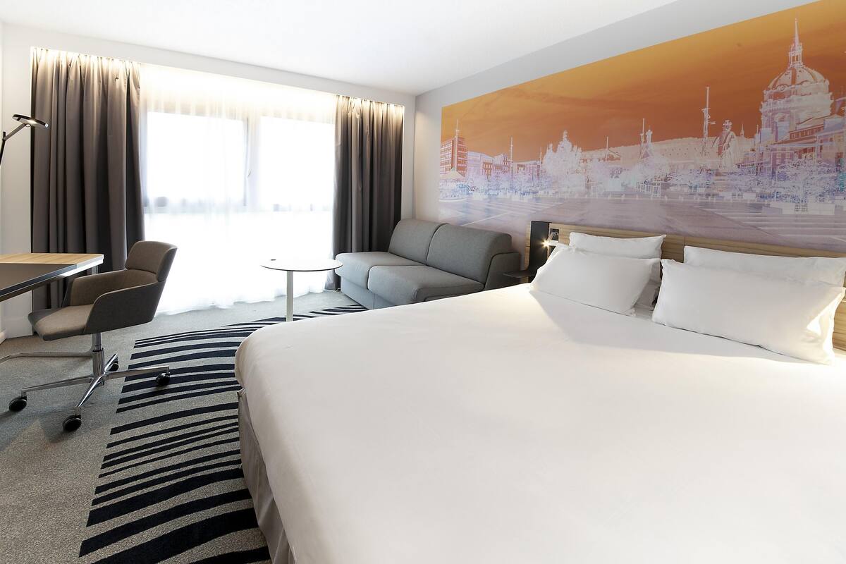 A hotel room with a large bed, a desk, a chair, a sofa, and a cityscape mural on the wall.
