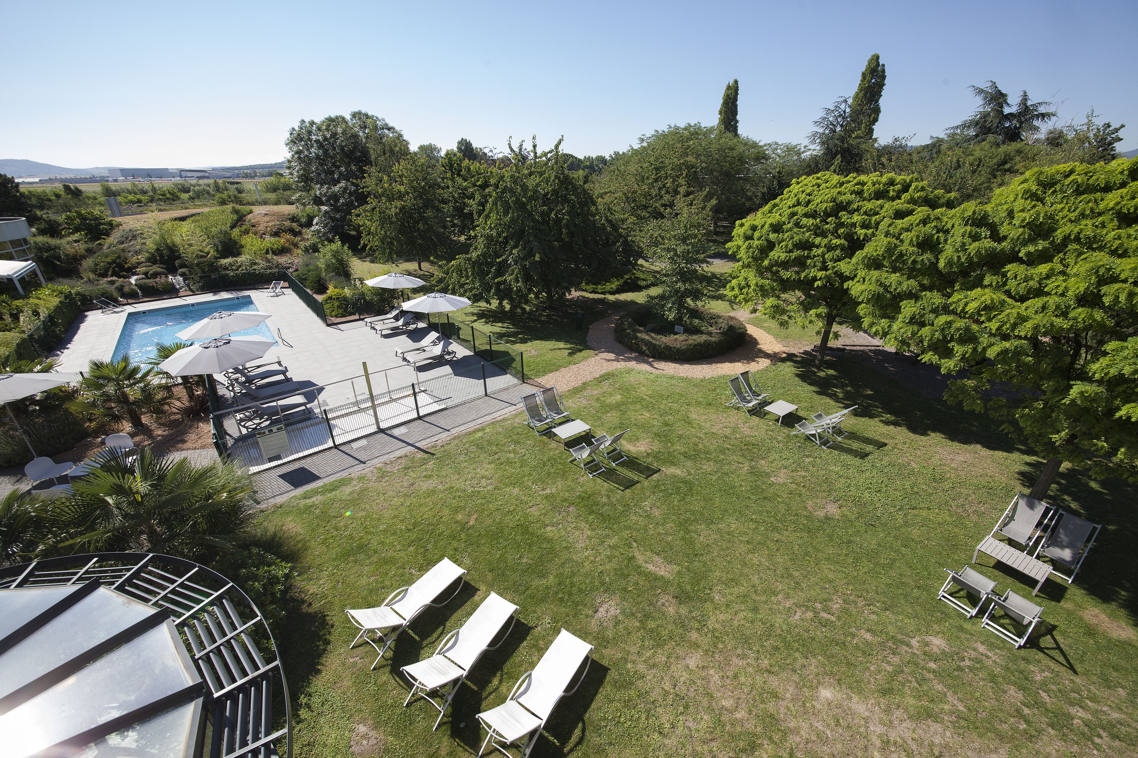 Zona de piscina con sillas de descanso y sombrillas, rodeada de árboles y un cielo despejado.