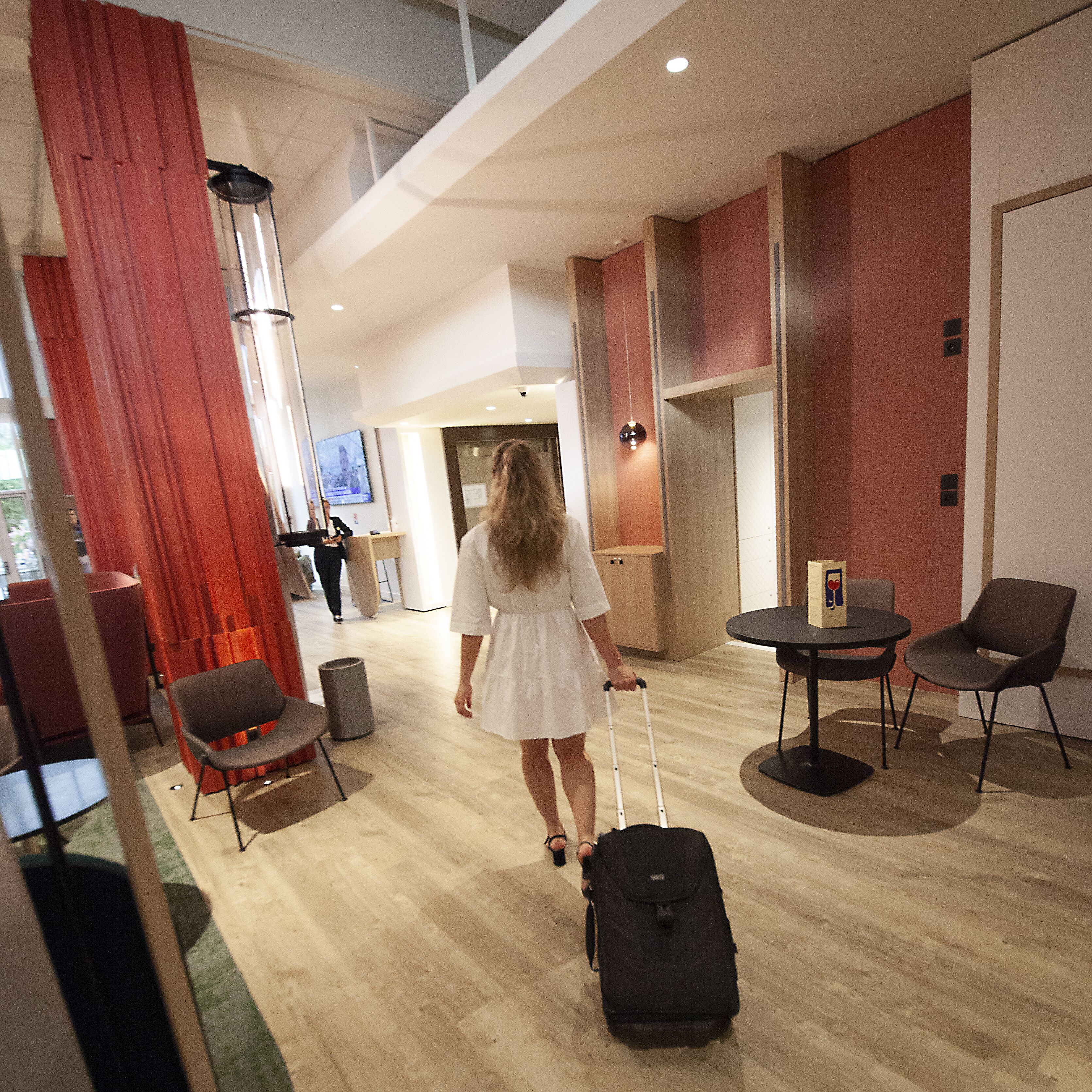A hotel lobby with a wooden floor, a red curtain, a black chair, a small table, and a person with a suitcase.