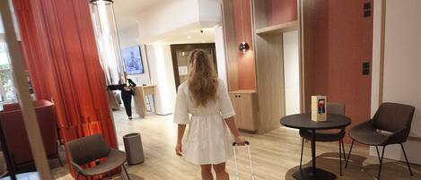 A hotel lobby with a wooden floor, a red curtain, a black chair, a small table, and a person with a suitcase.