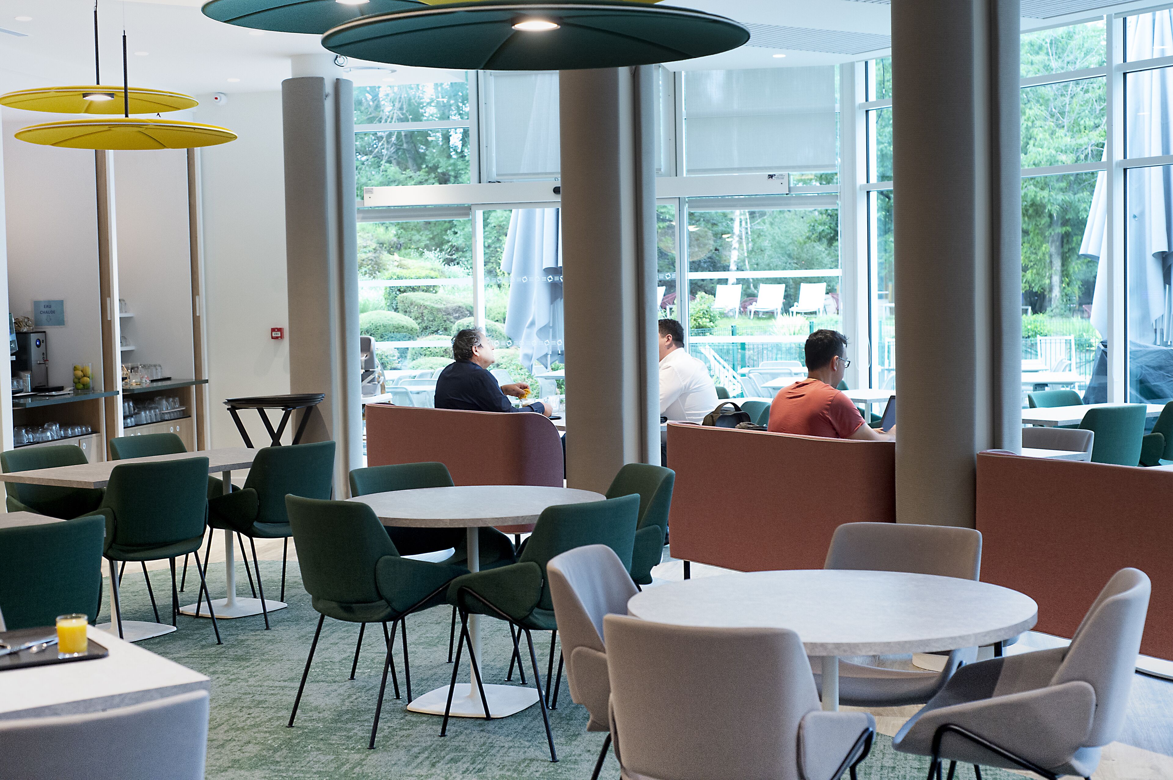 A modern dining area with round tables and green chairs, large windows, and a view of the outdoors.