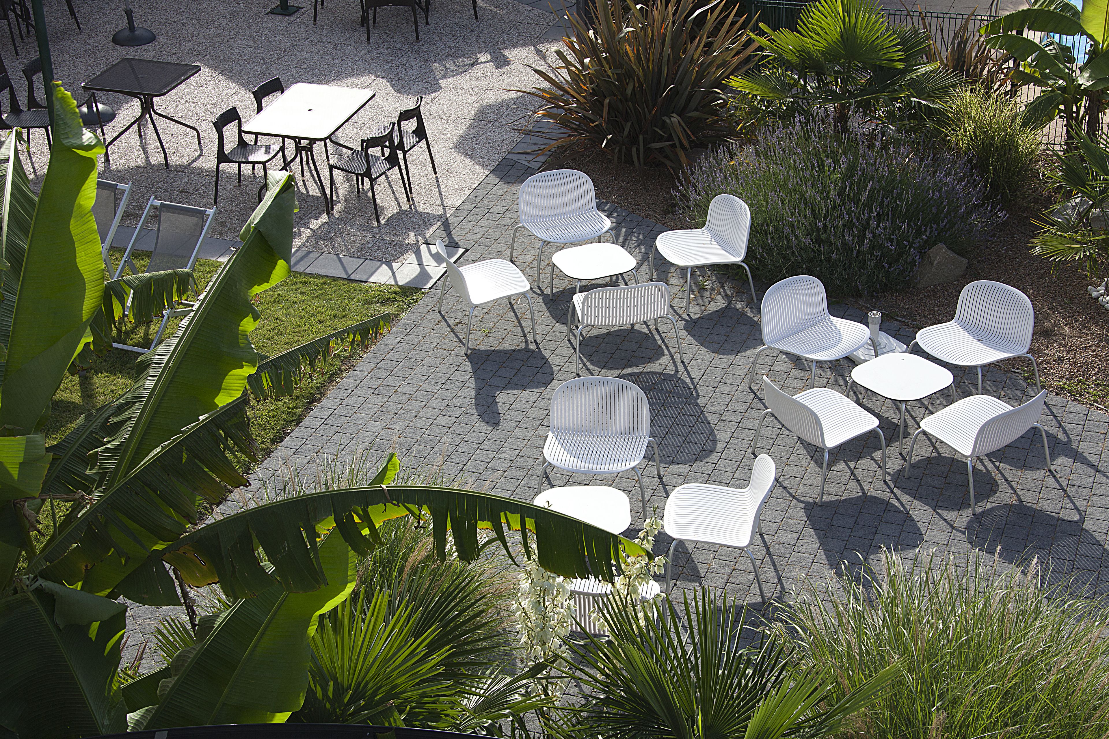 An outdoor patio area with white metal chairs and tables arranged on a paved surface, surrounded by lush green plants and palm trees.
