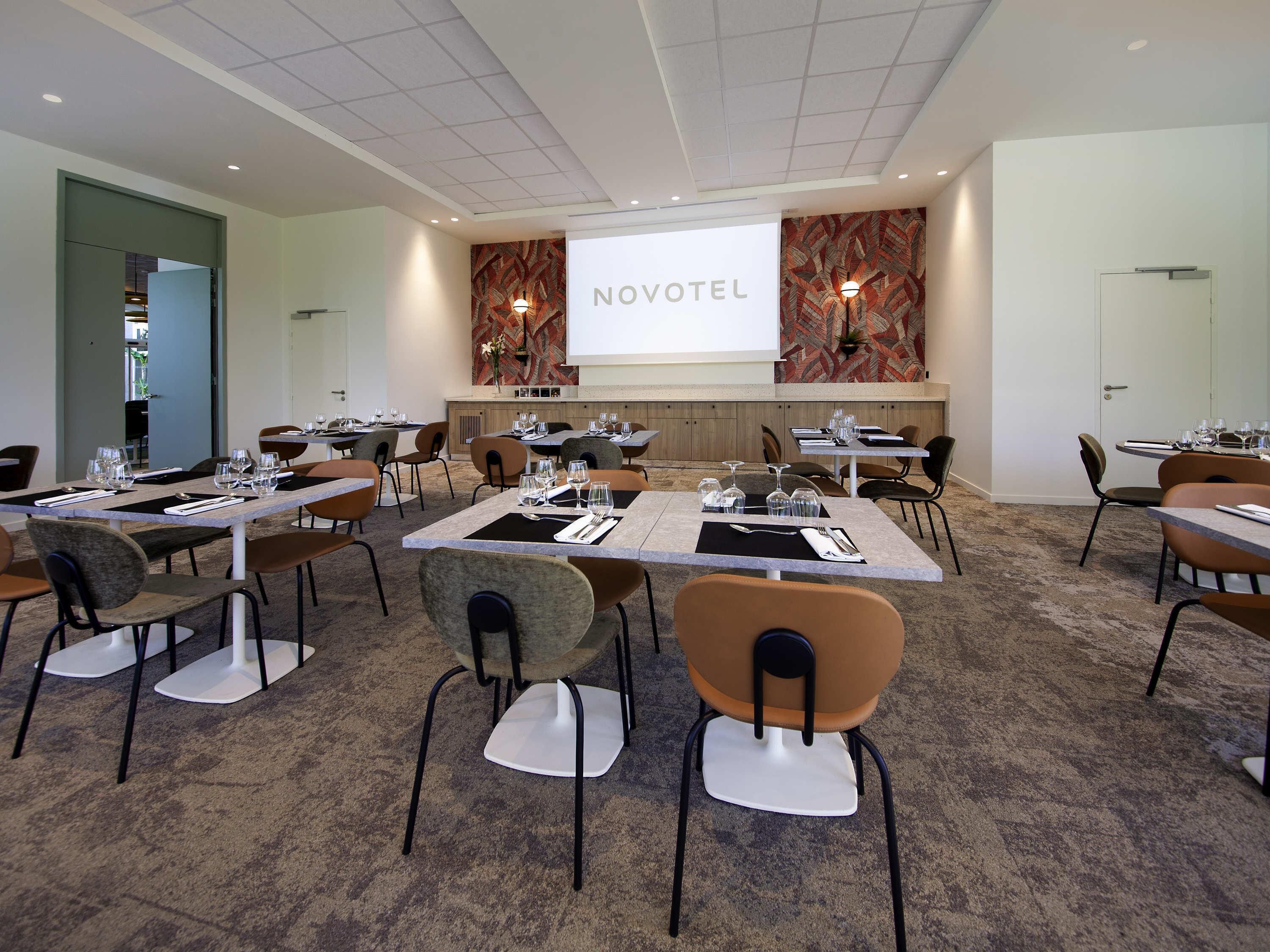 A conference room with tables set for a meeting, a projector screen displaying 'NOVOTEL', and a patterned wall.