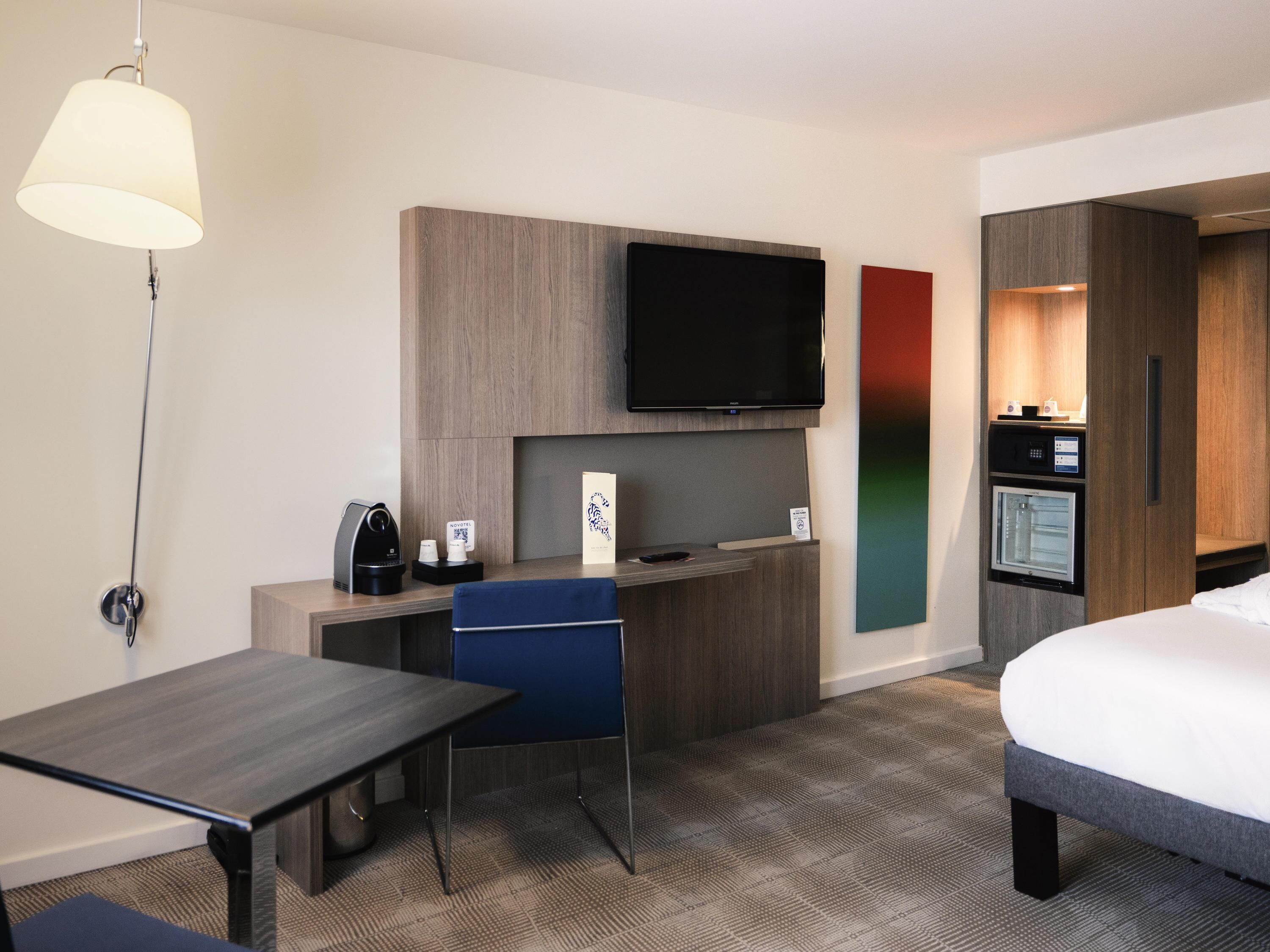 A hotel room with a bed, a desk with a chair, a television mounted on the wall, a minibar, and a coffee machine.