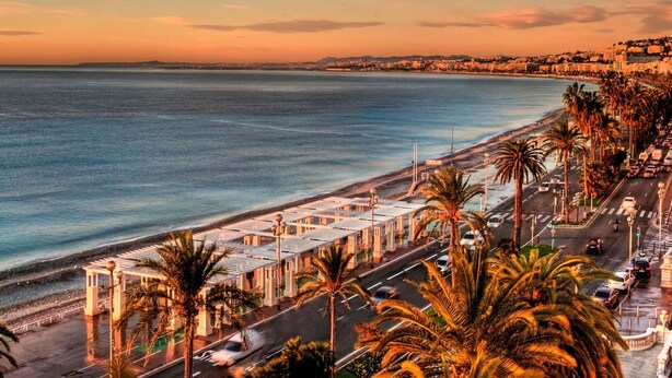 A coastal cityscape with a beach, palm trees, and a road with cars.