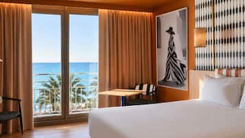 Habitación de hotel con cama, escritorio, silla, ventana grande con cortinas y vista al océano.