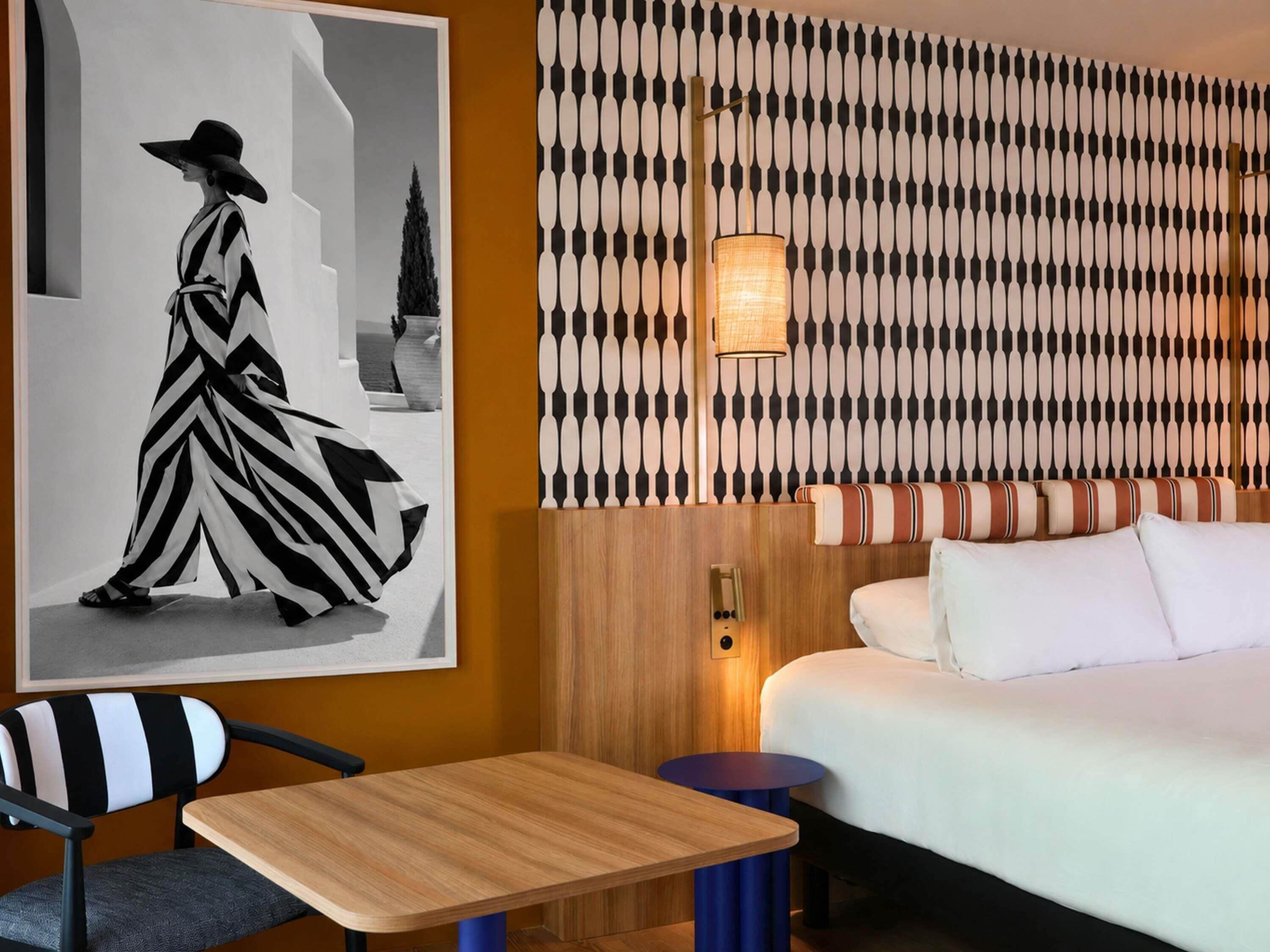 Habitación de hotel con cama, una mesita, una silla, pared con diseño decorativo y un cuadro en la pared.