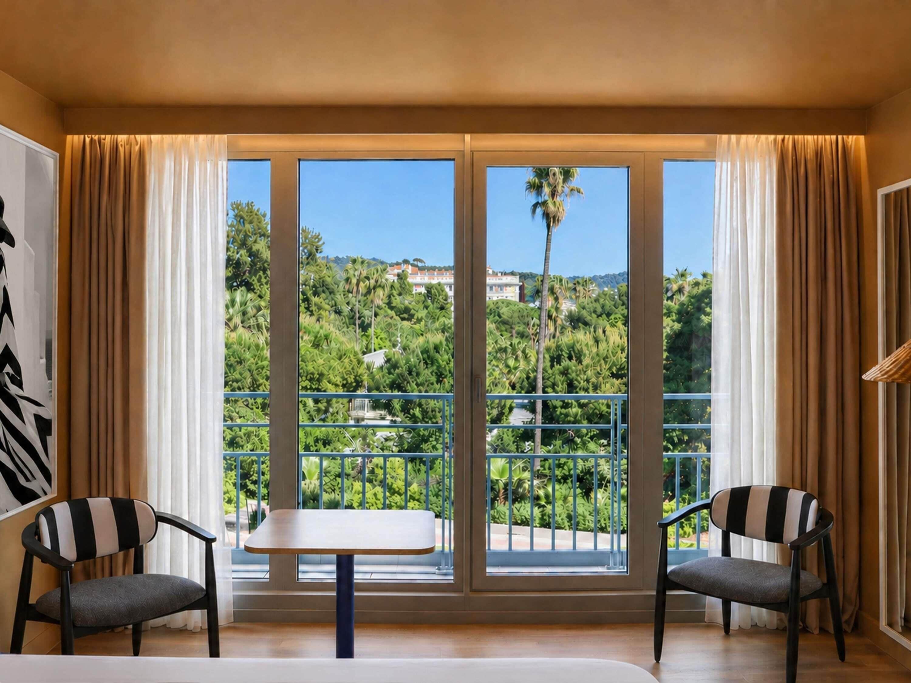 Une chambre avec un balcon, deux chaises, une table et une vue sur la verdure et les bâtiments.