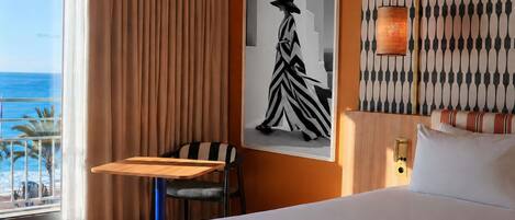 A hotel room with a bed, a desk, a chair, a large window with curtains, a framed picture on the wall, and a patterned wall design.