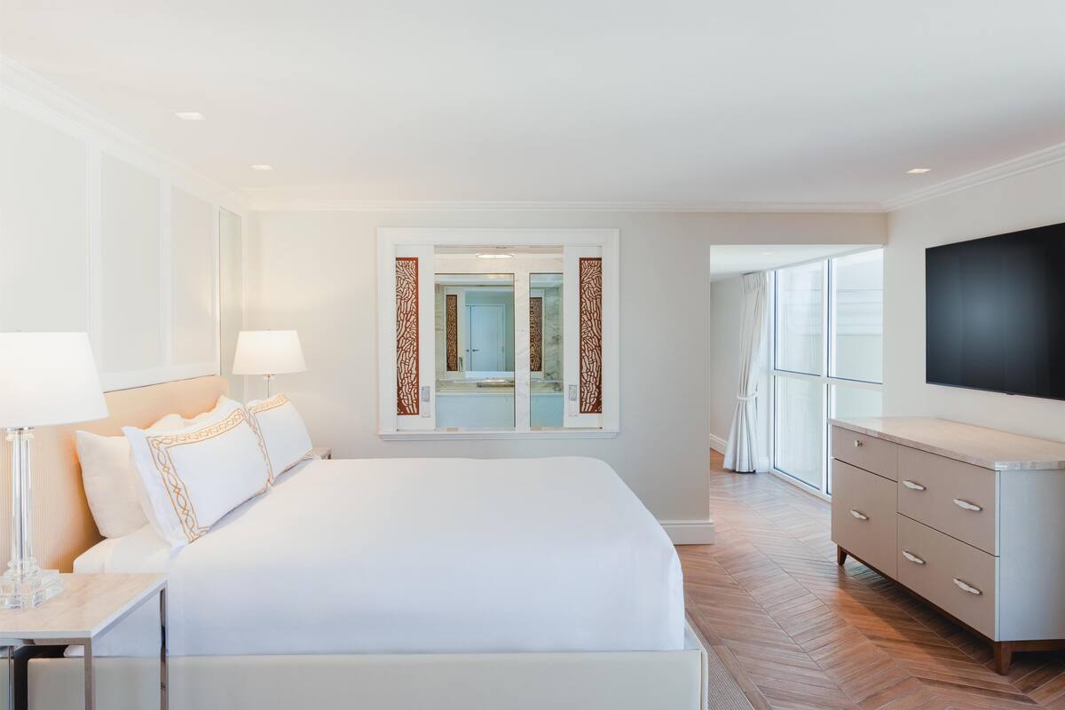 A hotel room with a large bed, bedside lamps, a TV, a dresser, and a window with curtains.