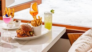A table with a meal, including a burger, fries, and drinks, by a window overlooking the ocean. - Surf & Sand Laguna Beach (Laguna Beach)