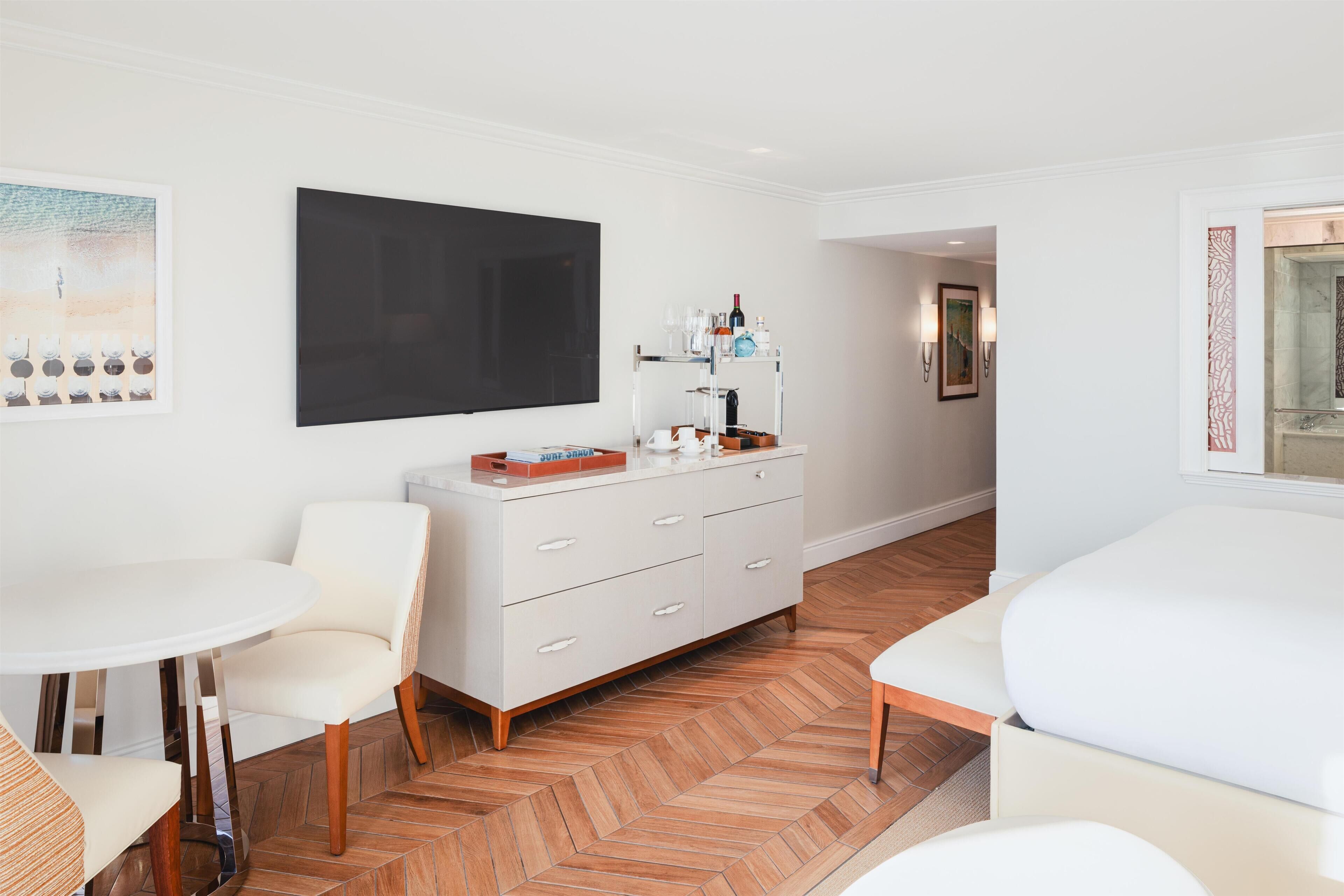 A hotel room with a white dresser, a round table, a sofa, a large black artwork on the wall, and a herringbone-patterned floor.