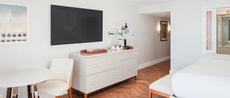 A hotel room with a white dresser, a round table, a sofa, a large black artwork on the wall, and a herringbone-patterned floor.