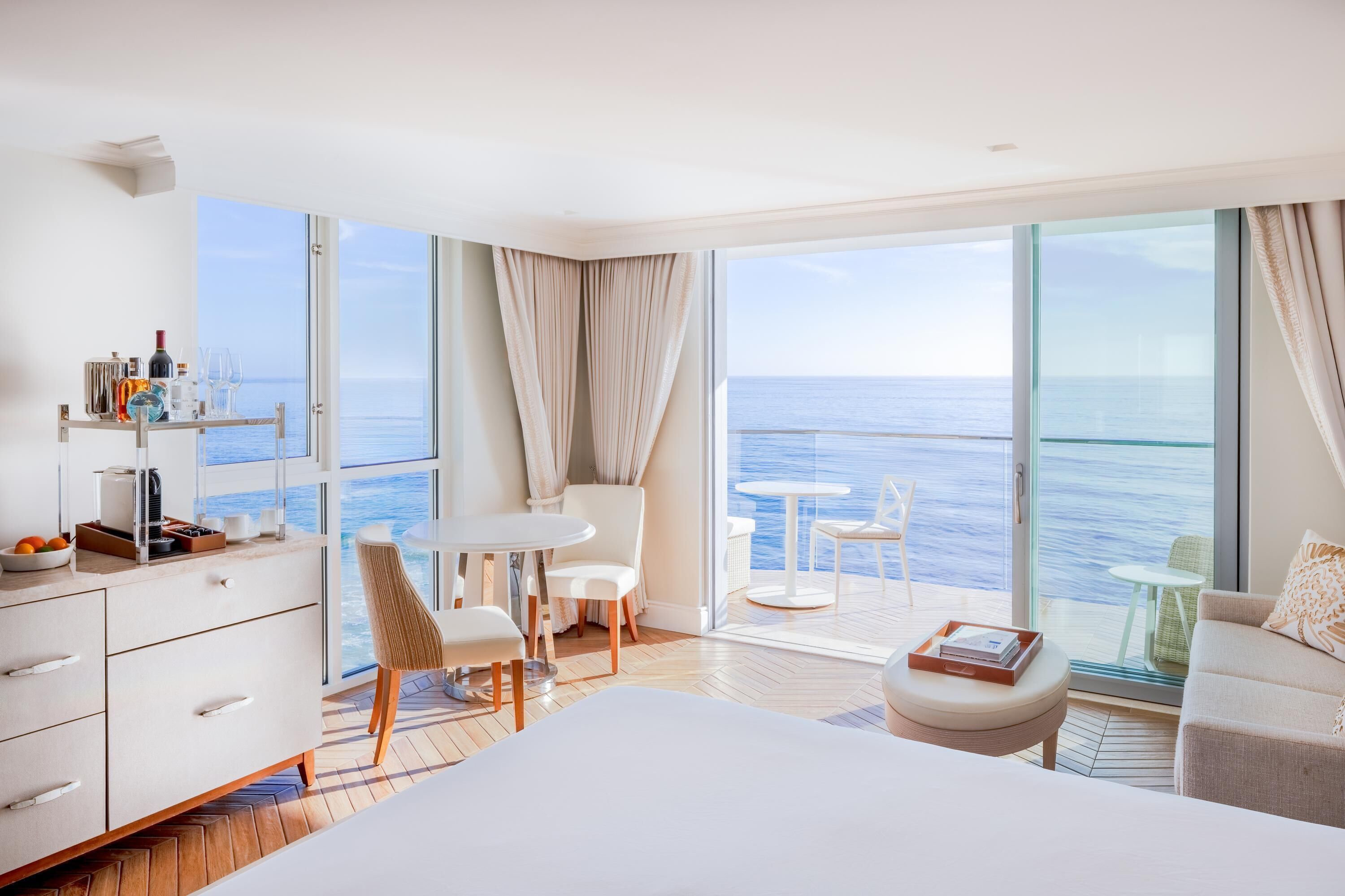A modern hotel room with a large bed, a desk, a chair, a small table, and a view of the sea.